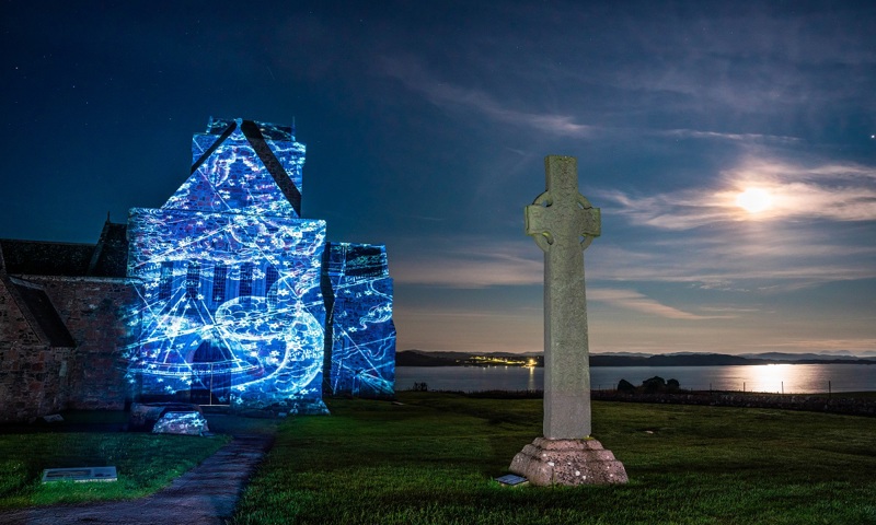 A Celtic cross standing in front of an abbey, upon which a dazzling blue pattern has been projected 