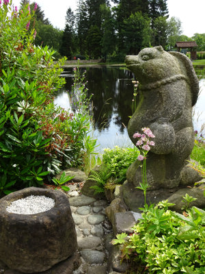 A stone panda sits facing away from a large pond, trees reflect in the water and a stone platform sits with rice in front of the panda