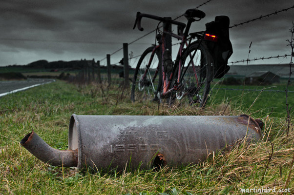 A bicycle leaning against a fence in Orkney next to a discarded car exhaust.