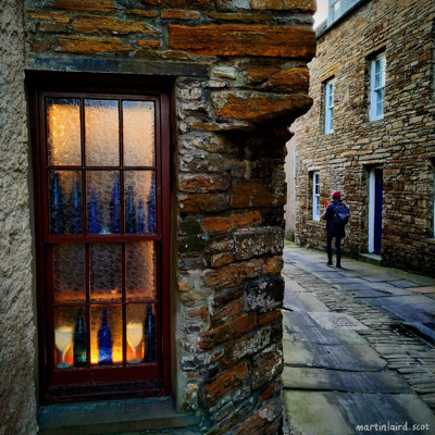 A warm, homely window in Stromness with bottles glowing through frosted glass. Outside black rubber coats the paving stones.