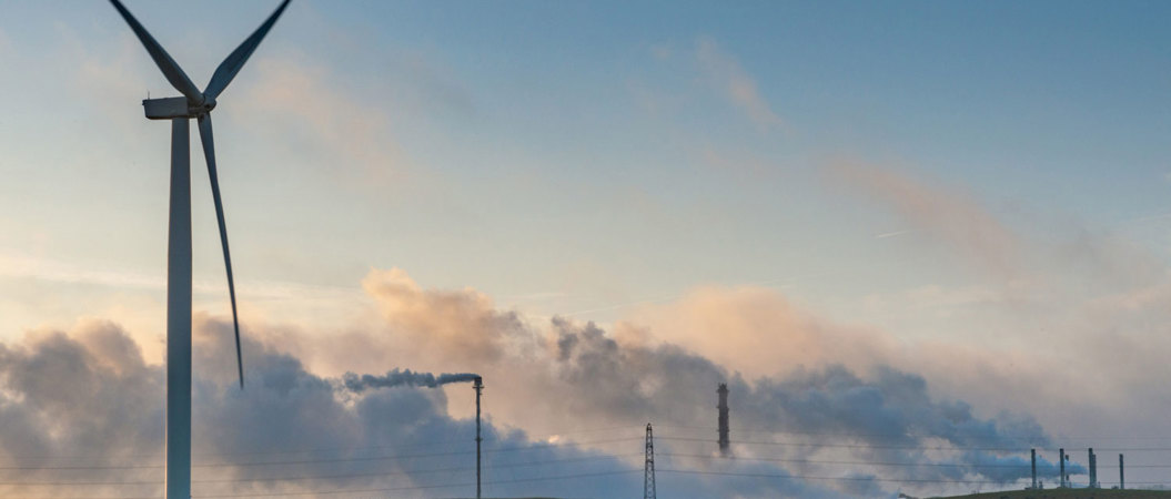 A wind turbine by a green field. In the distance, smoggy chimney can be seen chugging out fumes.