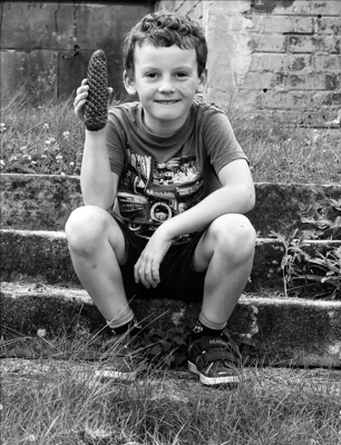 A young boy sits and smiles at the camera, proudly holding up a large pine cone