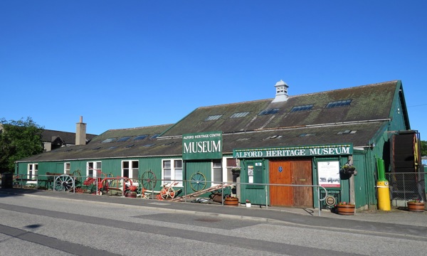 The exterior of a distinctive green, timbered auction building. As large, hand-painted signs indicate, it has been converted into a local heritage museum. Various agricultural artefacts, along with flower pots, are on display in front of the building.  