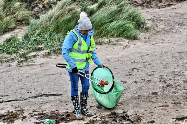 A woman in a hat and a high visibility jacket on a beach carrying in a bin bag