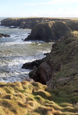 A heavily sunlit scenescape composed of coastal cliffs and coarse, light grass framing a view looking out over a pale sea