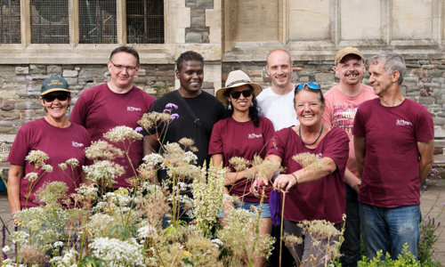 8 people smiling standing in front of a historic wall with some greenery in front of them