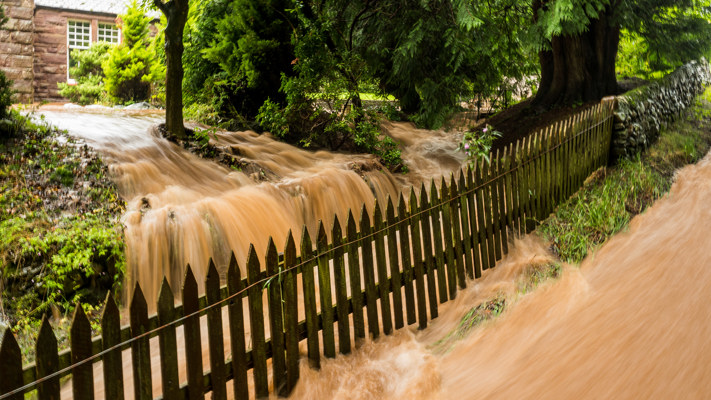 Flood water is coming past a house over a bank towards a stream. The foreground road is covered in fast flowing water.