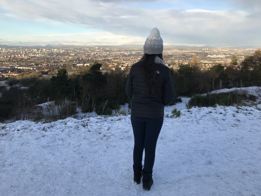 A girl stands on the top of a snow covered hill and looks out towards the city.