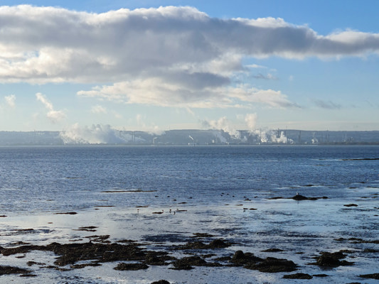 A landscape shot of the sea and a smokey, industrial chimneys on the other side of the water