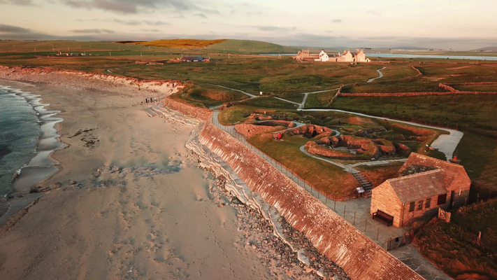 45º offset aerial image of Skara Brae Village facing encroaching waves, bathed in blood red sun light & encompassed by sea level background terrain
