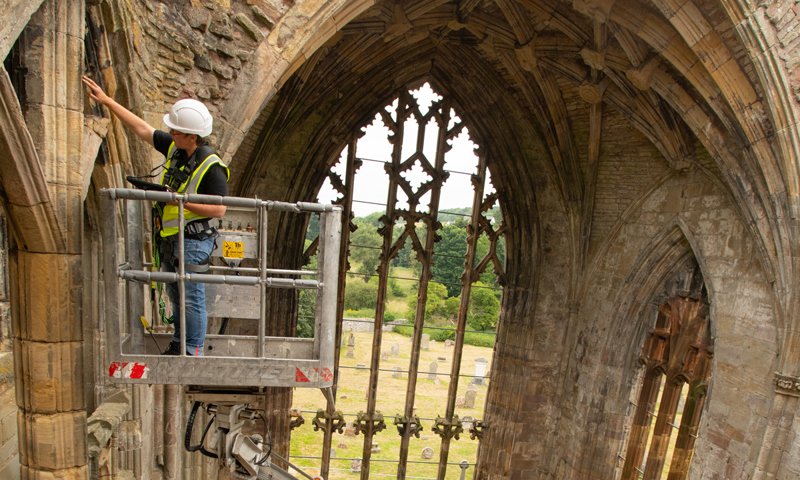 A person wearing personal protective equipment (PPE) while inspecting the stone archway of an abbey from the top of a cherry picker