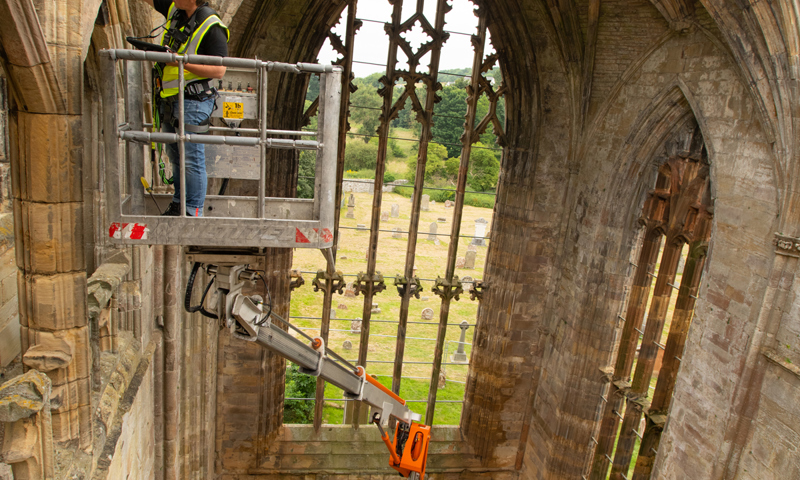 A person wearing personal protective equipment (PPE) while inspecting the stone archway of an abbey from the top of a cherry picker