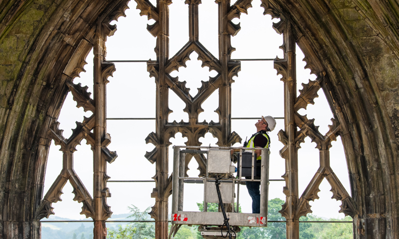 A person wearing personal protective equipment (PPE) while inspecting the stone archway of an abbey from the top of a cherry picker