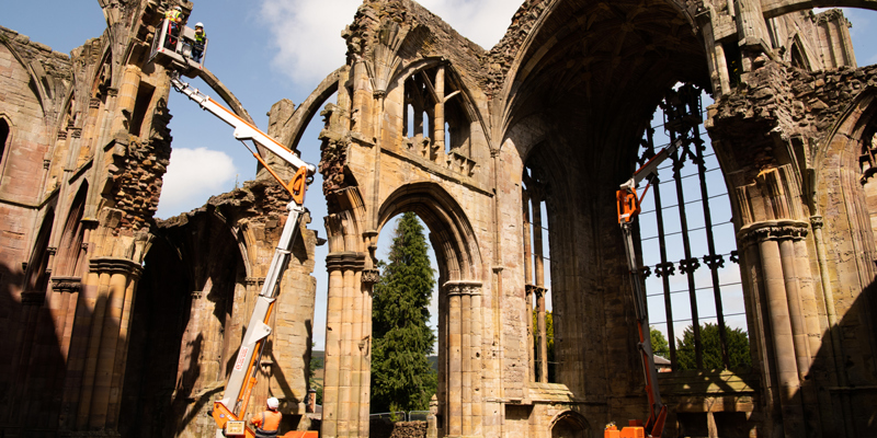 Engineers wearing personal protective equipment (PPE) while inspecting the masonry of an abbey from the top of two cherry pickers