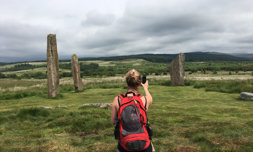 A lady wearing a rucksack takes a photo of remote standing stones for Monument Monitor using a smartphone 