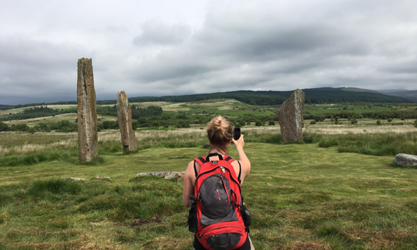 A lady wearing a rucksack takes a photo of remote standing stones for Monument Monitor using a smartphone 