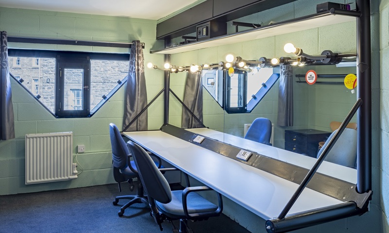 Inside a dressing room at a theatre. Hanging lights surround a large mirror behind a dressing table.