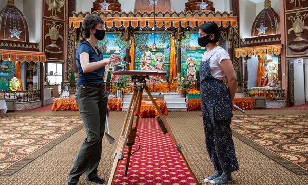Two people wearing facemasks stand facing each other, statues decorate the background of the room
