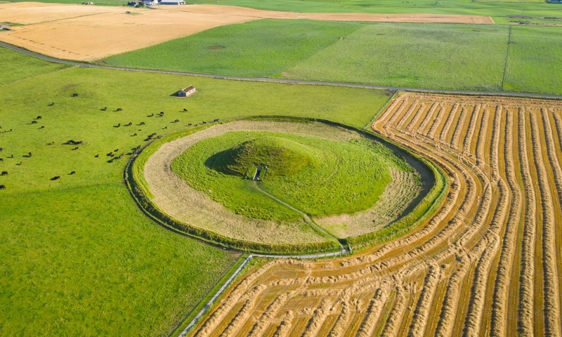 An aerial view of a grass-covered chambered cairn surrounded by a circular ditch and farmland