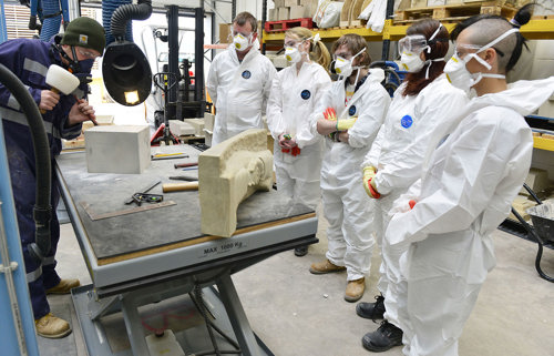 A group of five young people in protective clothing observe someone chiseling stone