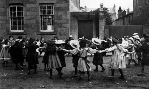 A black and white photo of girls in hats and dresses holding hands in a circle in a playground