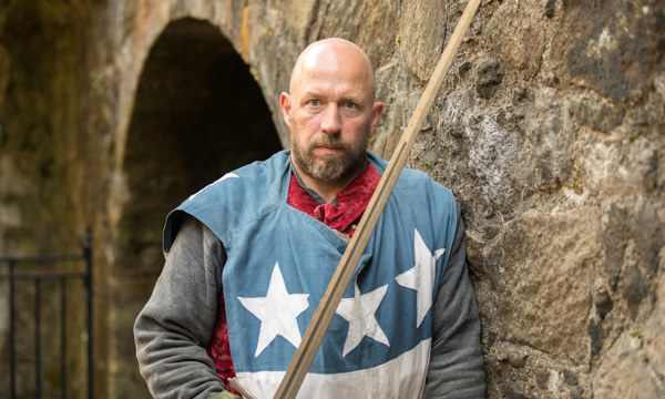 A Living History Character holding a sword leaning against a castle wall