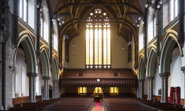 The interior of a large church building, A central aisle runs between banks of wooden pews towards a seating gallery and a large, ornate window, 
