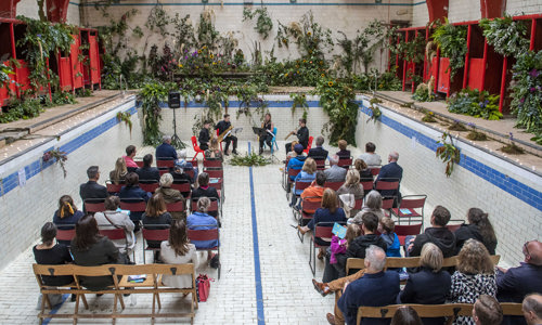 People sitting on seats in the empty pool at Govanhill baths with lots of flowers for the launch of Doors Open Day 2019
