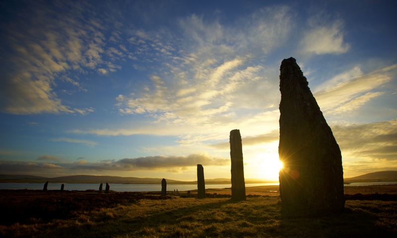 The setting sun behind three enormous standing stones, forming part of a large stone circle 