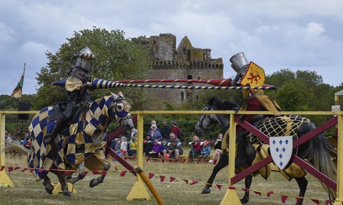 A landscape photo of two people in reenactment clothes dressed as knights sitting on horses. The knights are jousting in front of Caerlaverock Castle 