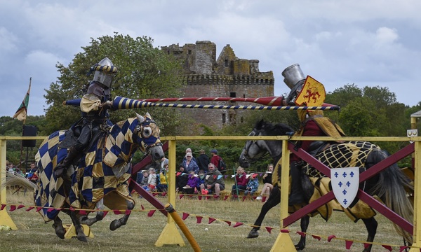 A landscape photo of two people in reenactment clothes dressed as knights sitting on horses. The knights are jousting in front of Caerlaverock Castle 