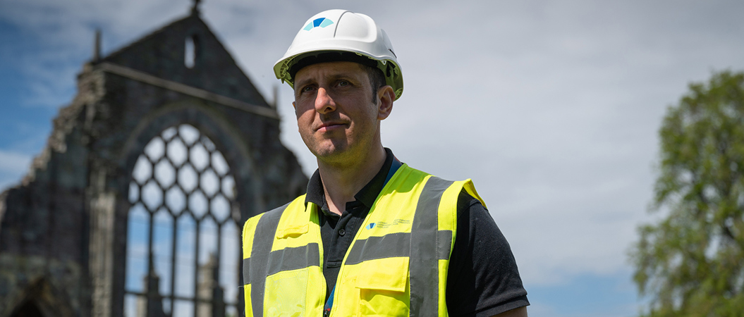 Male inspector wearing white safety helm and high-vis vest stands with Holyrood Abbey in the background on a sunny day
