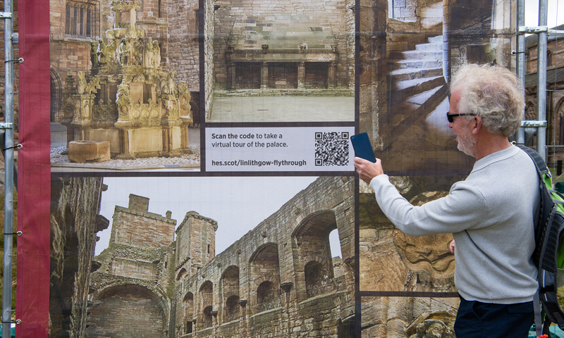 A man with white hair and wearing black sunglasses and a light blue jumper holds his phone up to a QR code on a board of various images of Linlithgow Palace.