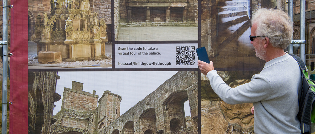 A man with white hair and wearing black sunglasses and a light blue jumper holds his phone up to a QR code on a board of various images of Linlithgow Palace.