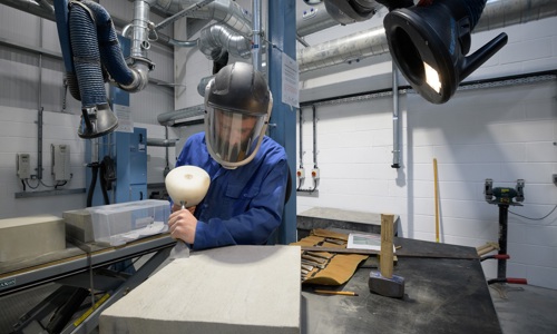 A person wearing protective gear chiseling a stone in a workshop.