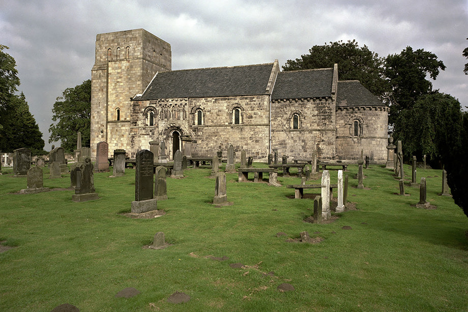 A small historic stone church surrounded by grass and trees, with gravestones in the graveyard