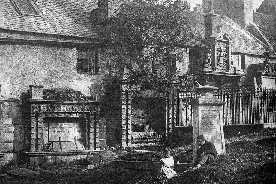 Two people sitting beneath a gravestone in a graveyard