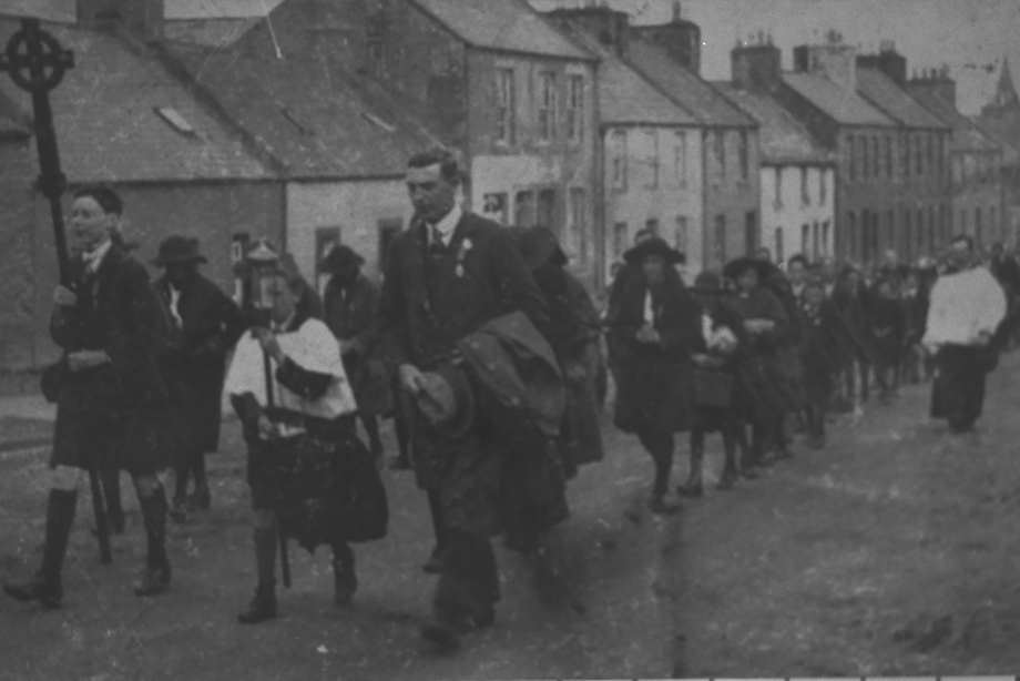 A large procession of people following a man holding a cross through the streets