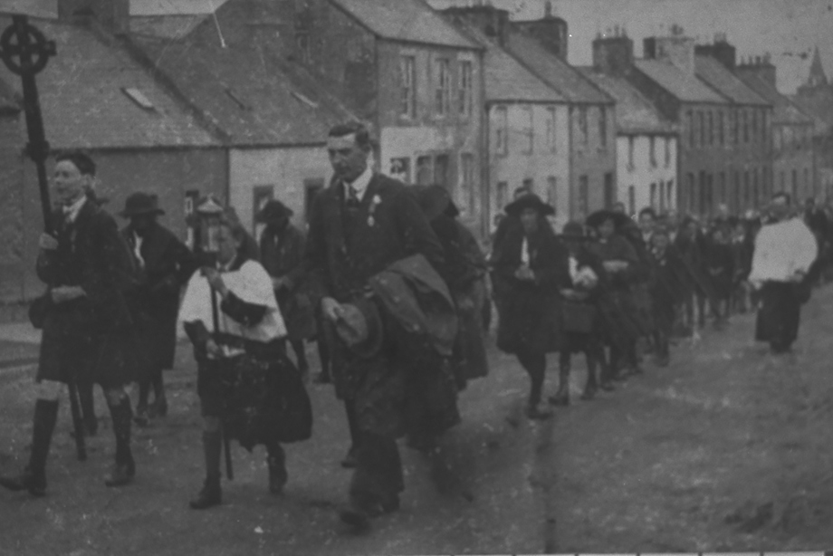 A large procession of people following a man holding a cross through the streets