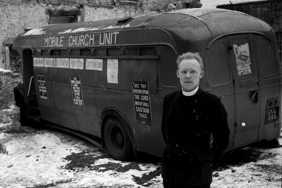 A reverend wearing his uniform, standing outside a bus with religious posters on the side