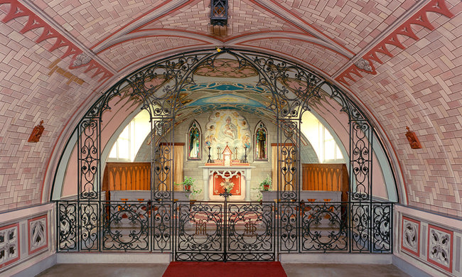 A rounded chapel building with pink, brick-effect walls and ironwork fencing leading into the altar 