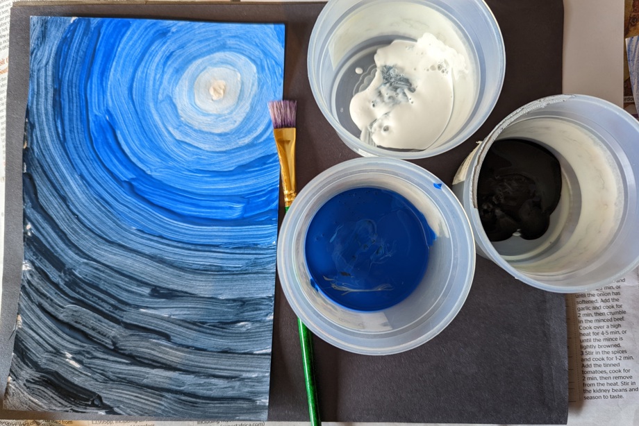 Blue, black and white paint pots and paintbrush next to a painting of moonlit sky.
