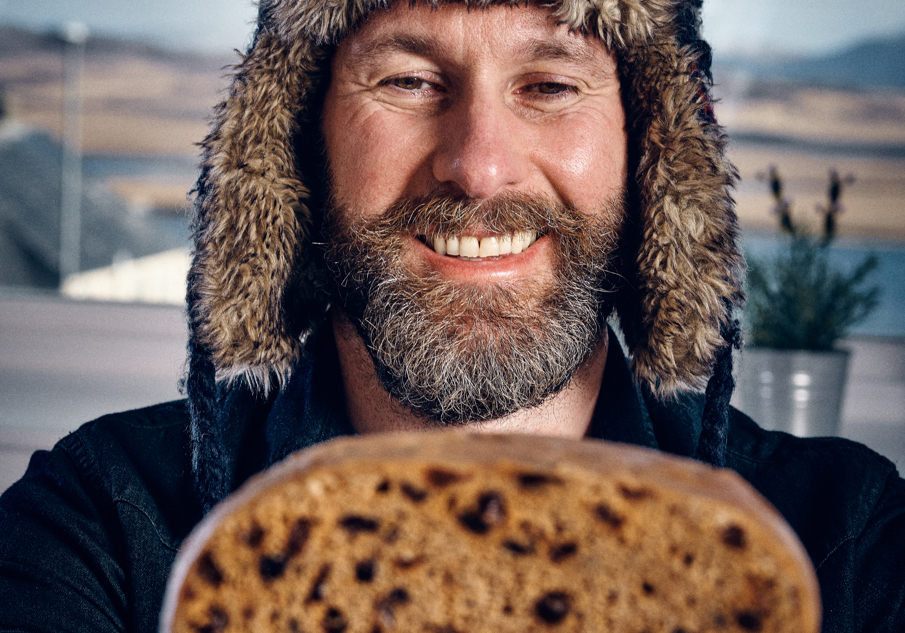 Smiling man with a salt and pepper beard, wearing an aviator hat, holds up to the camera a clootie dumpling cut in half.
