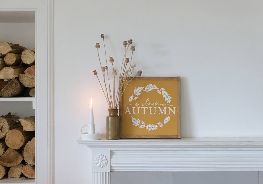 A candle, a brown vase with dried flowers, and a picture frame with the words "Welcome Autumn" written on them sit on top a white, fire mantlepiece.