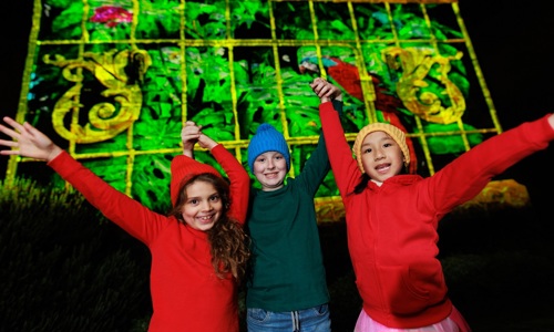 Three children pose excitedly in front of a brightly illuminated building inside Edinburgh Castle