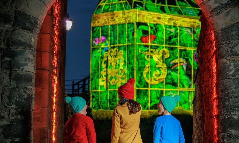 Three children in brightly coloured outfits walking through an archway at Edinburgh Castle. A chapel illuminated with projections can be seen in the background. 