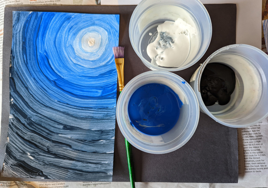 From from above three plastic cups, containing white, black and blue paint at the bottom. Beside them is a green paint brush and a small painting of a moon whose light spreads out in a circular pattern from white to blue to black.