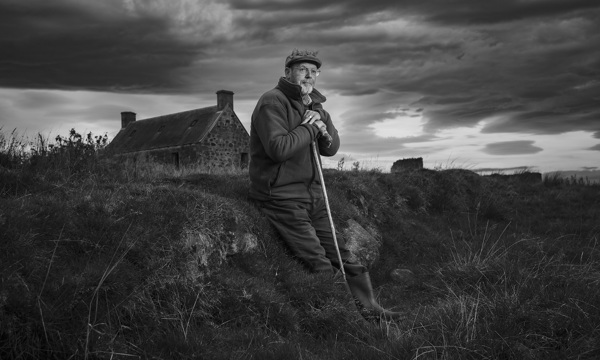 Black and white photo of an older man leaning on a shepherd's crook outside a stone cottage.