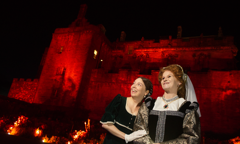 Two costumed reenactors in a 1920s style dress and a medieval style dress in front of Stirling Castle which is illuminated with red floodlights
