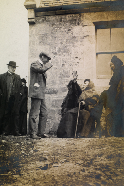 People standing around a cloaked, seated woman holding one hand in the air and another on a walking stick, outside a stone building. Image reference number:  DP_102555